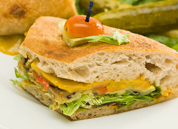 A close-up of a delicious vegetarian sandwich with layers of lettuce, tomatoes, cheese, and herbs, garnished with a cherry tomato and pickle on top.