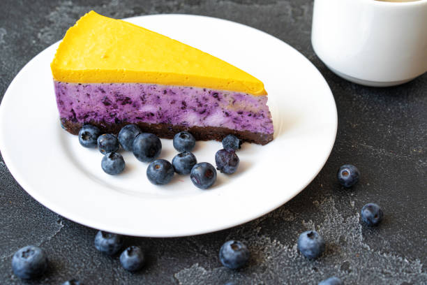 A slice of two-layered cheesecake with a yellow top and purple bottom on a white plate, surrounded by fresh blueberries.