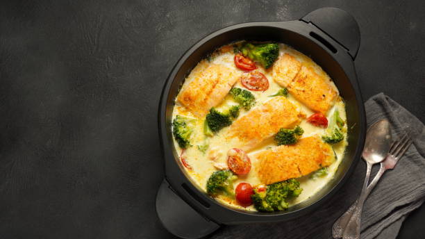 A creamy dish with salmon fillets surrounded by broccoli and cherry tomatoes in a dark pot on a black background.