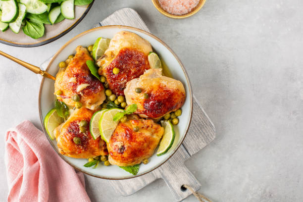 A plate of golden-brown chicken thighs garnished with lime slices and green peas, served on a wooden board with a side of sliced cucumbers and sea salt.