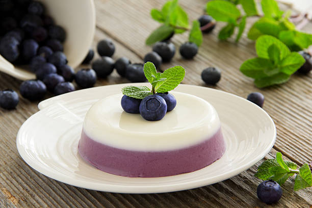 A creamy layered dessert topped with fresh blueberries and mint leaves, served on a white plate with spilled blueberries around it.