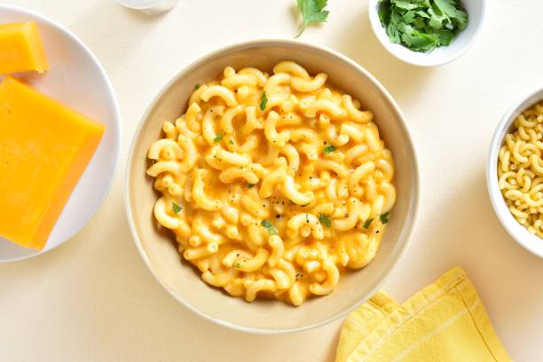 Bowl of creamy macaroni and cheese with fresh herbs, surrounded by cheese and uncooked pasta.