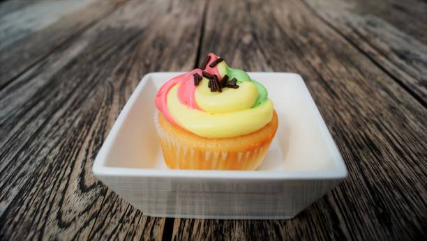 A colorful cupcake with pink, yellow, and green frosting, topped with chocolate sprinkles, sitting in a white dish on a wooden table.