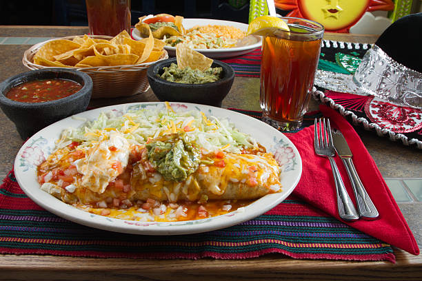 A colorful plate of enchiladas topped with cheese, lettuce, and guacamole, served with sides of salsa, tortilla chips, and a drink.