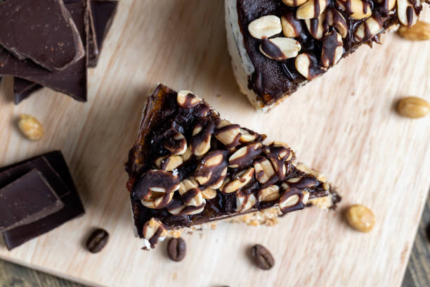 A slice of chocolate dessert topped with peanuts on a wooden board, accompanied by chocolate squares and coffee beans.