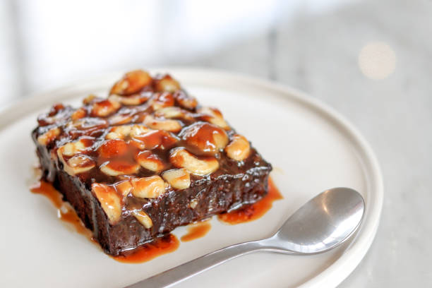 A delicious chocolate brownie topped with nuts and caramel sauce served on a white plate with a spoon.