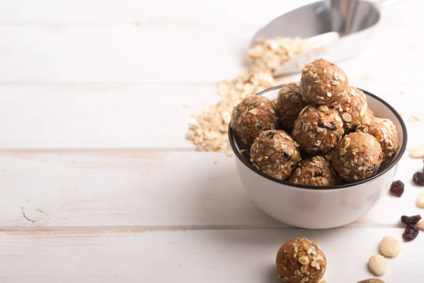 A bowl of healthy snack energy balls made from oats, nuts, and dried fruits, with some scattered around on a light wooden surface.