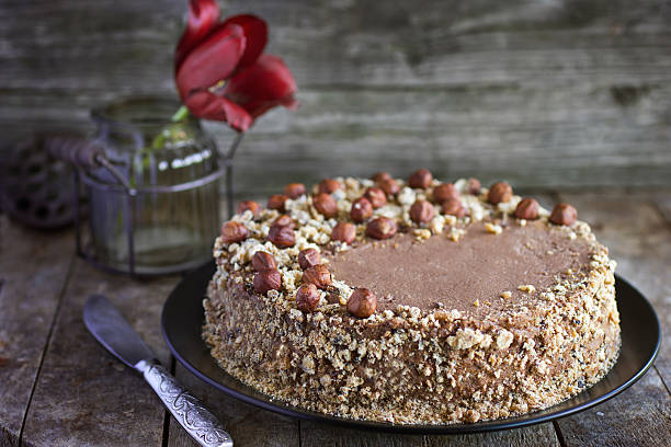 A round chocolate cake topped with hazelnuts on a dark plate, with a vintage knife and a flower in the background.