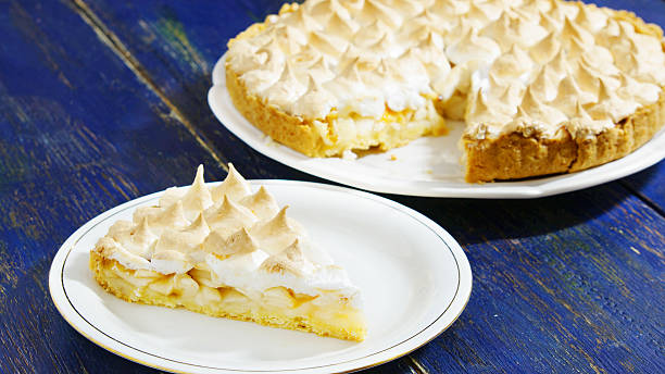 A slice of lemon meringue pie on a plate next to a whole pie on a white platter, set against a blue wooden background.