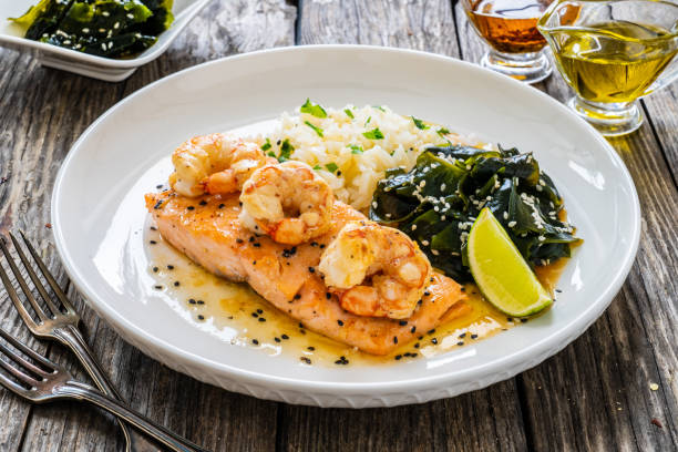 A plate of salmon topped with shrimp, served with rice and sautéed greens, garnished with lime.