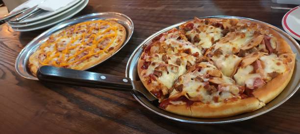 Two pizzas on metal serving trays on a wooden table, one topped with cheese and the other with a variety of meats.