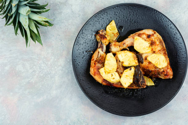 Grilled chicken pieces topped with pineapples on a black plate, with a pineapple in the background.