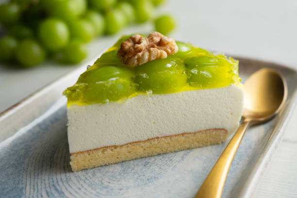 A slice of creamy dessert topped with green gelatin and a walnut on a plate, with green grapes in the background.