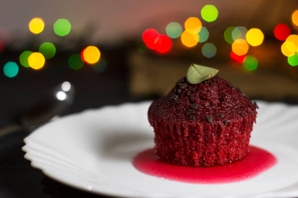 A single red velvet cupcake on a white plate with a berry sauce and chocolate sprinkles, set against a backdrop of colorful bokeh lights.