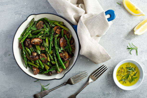 A bowl of sautéed asparagus and mushrooms on a grey surface with lemon wedges and olive oil on the side.