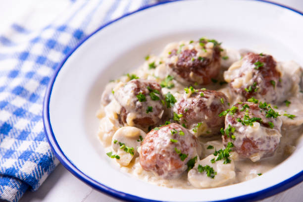 A plate of meatballs in a creamy sauce topped with chopped parsley, served with mushrooms, alongside a blue and white checkered cloth.