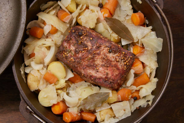 A hearty meal featuring a piece of meat surrounded by vegetables, including carrots, cabbage, and potatoes, in a rustic pot.