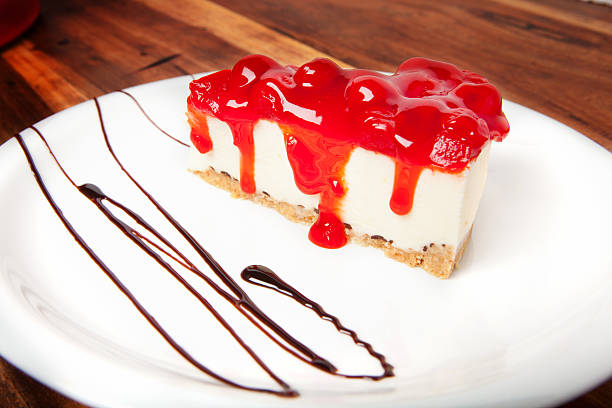 A slice of cheesecake topped with a glossy red cherry sauce drizzled on a white plate with a chocolate sauce decoration.