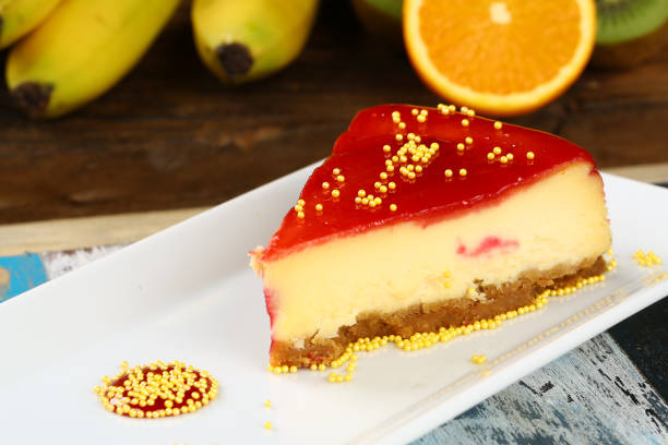 A slice of cheesecake with a red jelly topping and decorative sprinkles on a white plate, with bananas and an orange in the background.