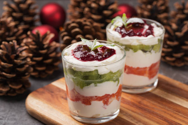 Two dessert glasses filled with layers of cream, fruit, and jelly, garnished with mint leaves, set against a backdrop of pine cones and Christmas ornaments.