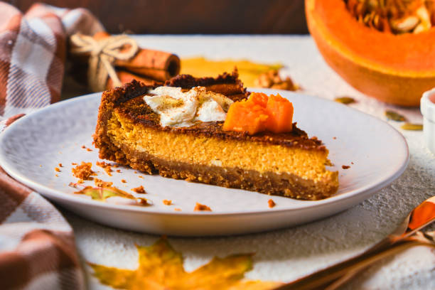 A slice of pumpkin cheesecake on a white plate, garnished with whipped cream and pumpkin pieces, surrounded by autumn leaves and spices.