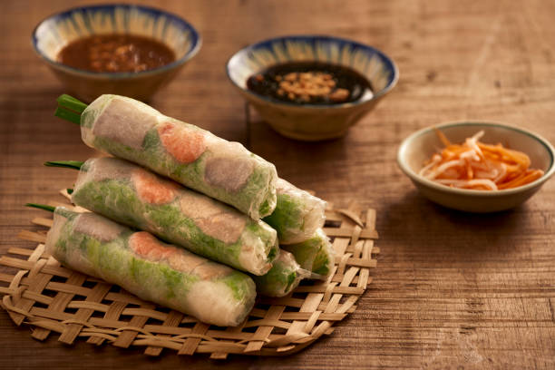 Freshly made spring rolls arranged on a woven bamboo mat, with side dishes in the background.