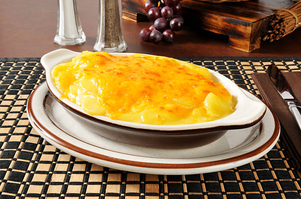 A serving of cheesy potato gratin in a white dish on a plate, resting on a woven mat with a wooden backdrop and grapes in the background.