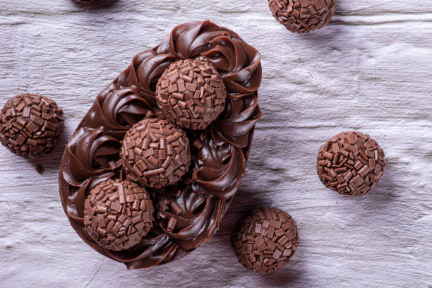 A chocolate dessert featuring an intricately decorated chocolate egg topped with chocolate truffles and decorative sprinkles, set against a light wooden background.