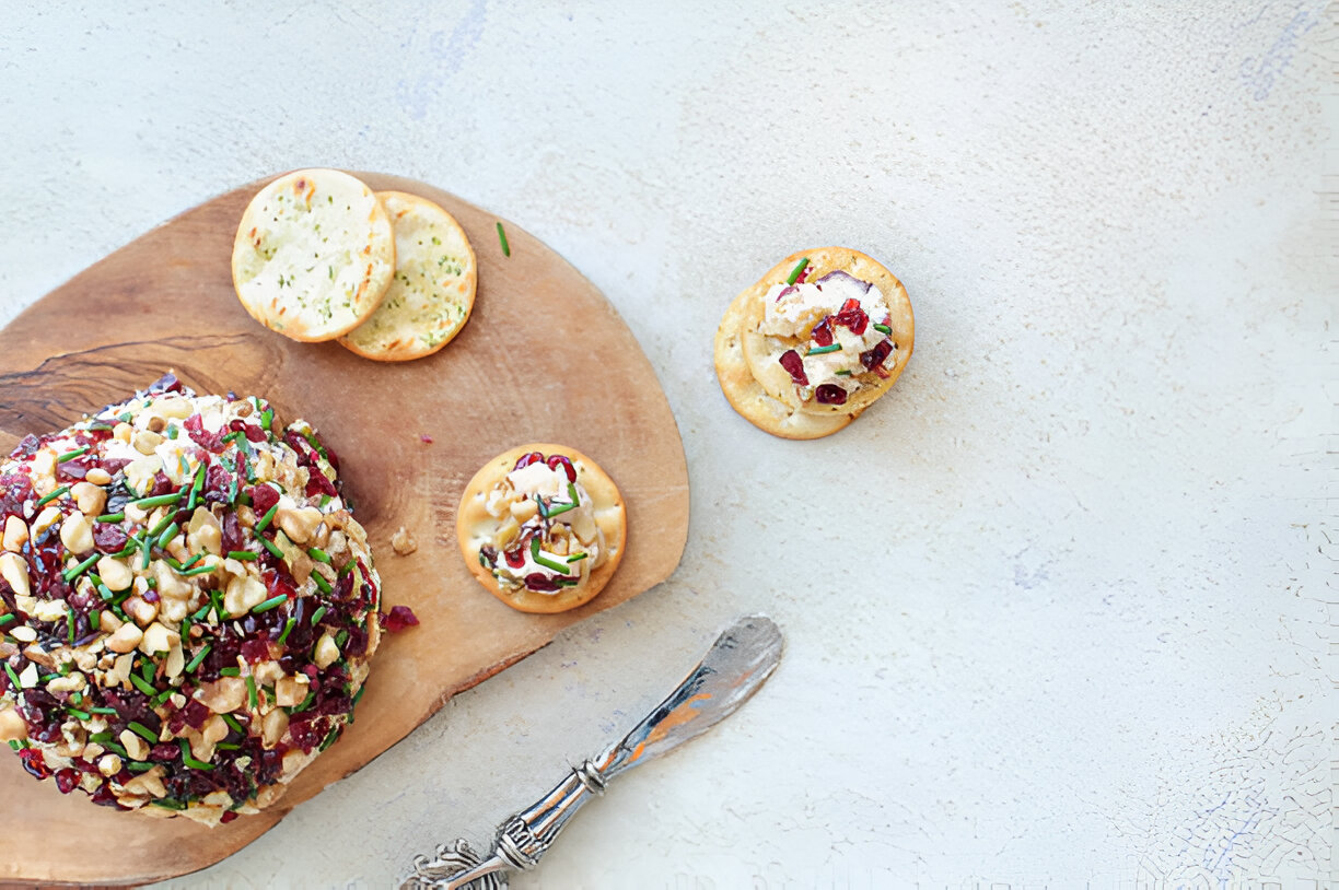 A wooden board with a cheese ball topped with nuts and cranberries, accompanied by crackers on a light background.