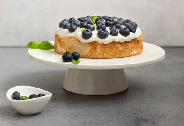 A delicious blueberry cake topped with fresh blueberries and mint leaves, displayed on a white cake stand.