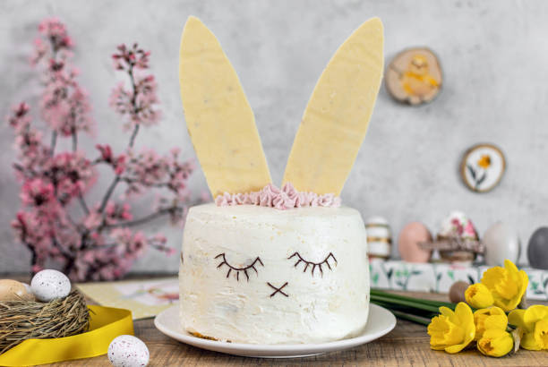 A creatively decorated cake designed to look like a bunny, with white frosting, chocolate facial features, and white chocolate bunny ears, surrounded by flowers and Easter decorations.