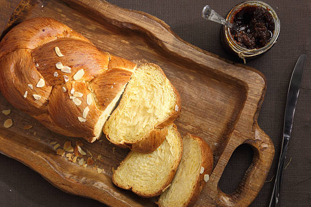 A sliced loaf of braided bread on a wooden serving tray with a jar of jam and a knife.