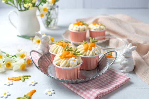 Colorful cupcakes decorated with carrot and flower designs on a silver tray amidst spring flowers and cute bunny figurines.