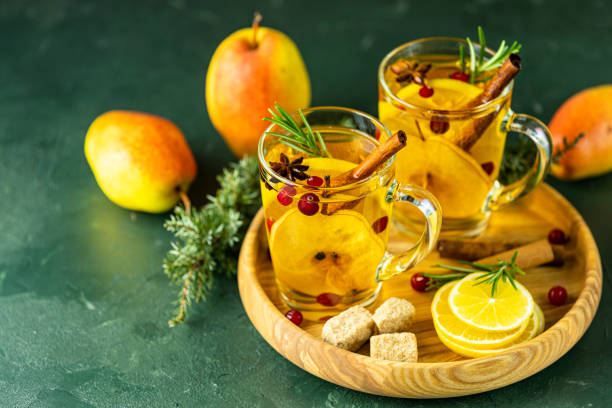 Two glass mugs filled with a warm spiced drink, garnished with apple slices, cinnamon sticks, and berries, alongside fresh apples and lemon on a wooden tray.