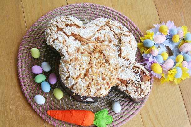 A creatively decorated cake shaped like a bunny, surrounded by colorful Easter eggs and a felt carrot, placed on a round woven mat.