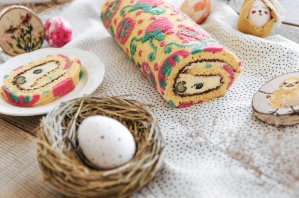 A colorful rolled cake with decorative patterns, sliced to reveal a cream filling, placed among Easter-themed items including a nest with an egg and decorated eggs.