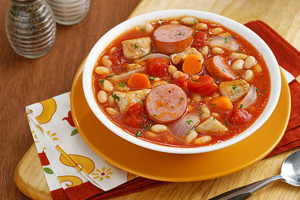 A hearty bowl of bean and sausage soup with carrots and tomatoes, served in a white bowl on an orange plate.