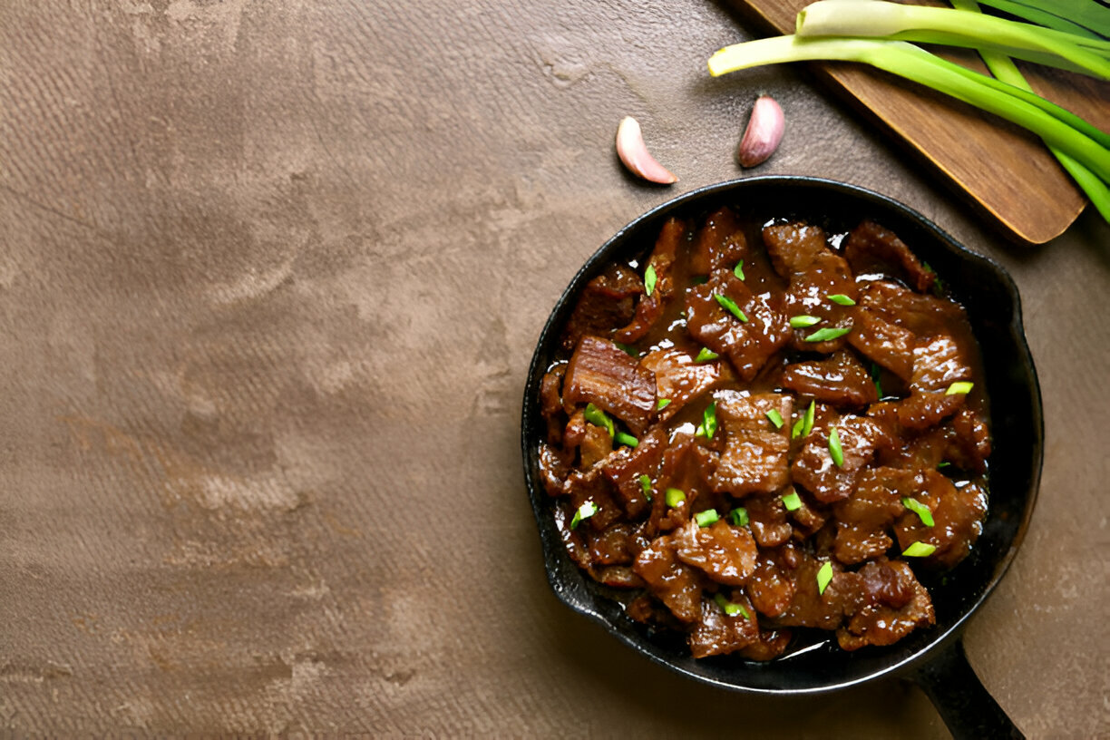 A cast iron skillet filled with a savory beef dish garnished with green onions, accompanied by garlic cloves and green onions on the side.