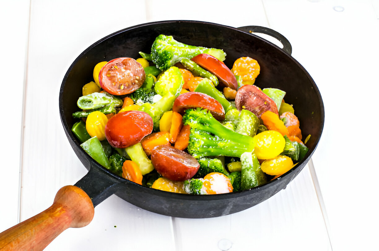 A black skillet filled with a colorful mix of frozen vegetables including broccoli, green peppers, and cherry tomatoes on a white wooden surface.