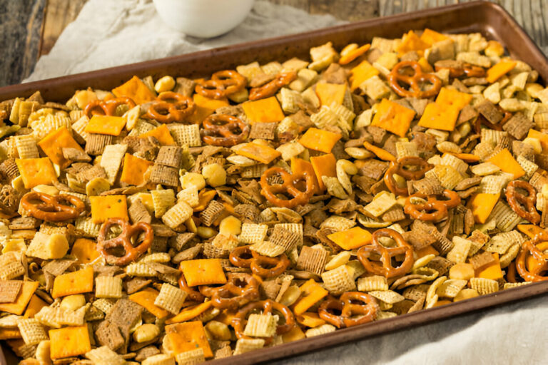 A close-up of a baking sheet filled with a colorful mix of cereal, pretzels, and cheese crackers.