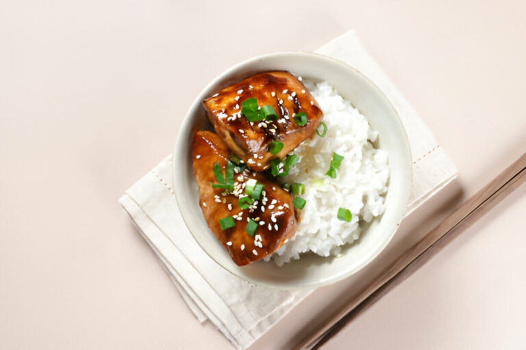 A bowl of rice topped with glazed chicken and green onions, served with chopsticks