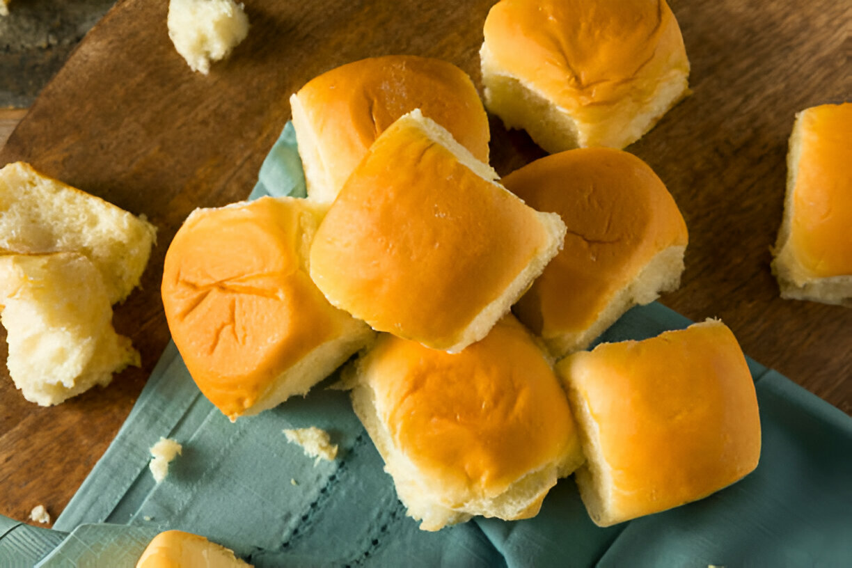 A pile of soft, golden dinner rolls on a wooden surface with a teal cloth.