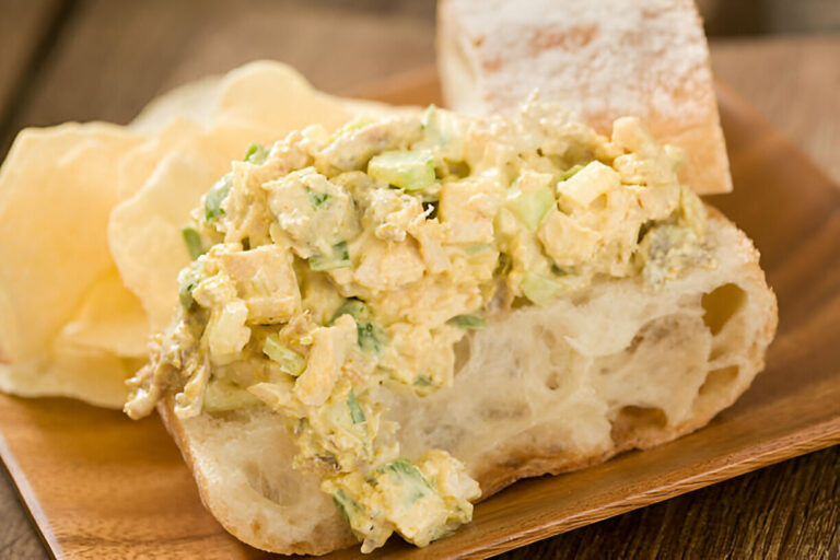 A slice of bread topped with a creamy chicken salad, served with potato chips and a piece of bread on a wooden plate.