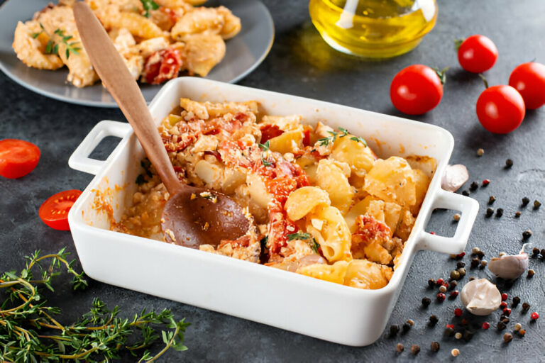 Dish of creamy pasta with tomatoes and herbs, served in a white casserole with a wooden spoon, surrounded by cherry tomatoes and spices.