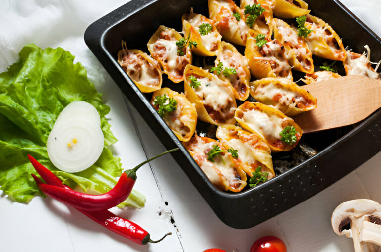 A baking dish filled with cheesy stuffed pasta shells topped with parsley, surrounded by fresh vegetables including lettuce, onion, and red chili peppers.