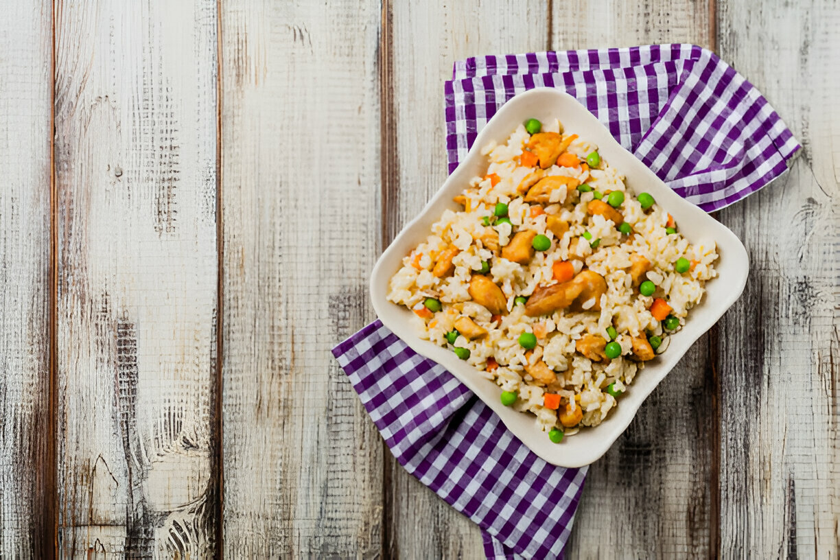 A bowl of chicken fried rice with green peas and carrots, served on a purple checkered napkin.