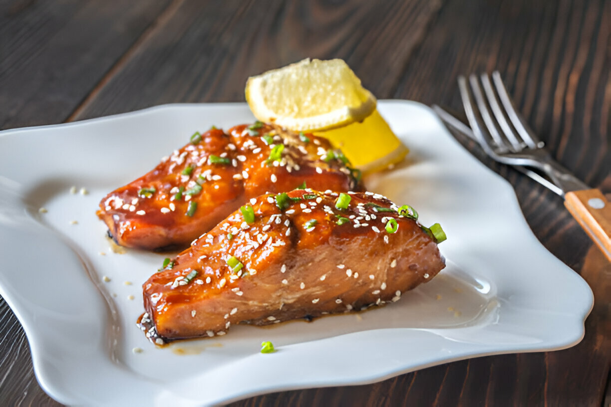 Two pieces of glazed fish garnished with sliced green onions and sesame seeds on a white plate, with a slice of lemon