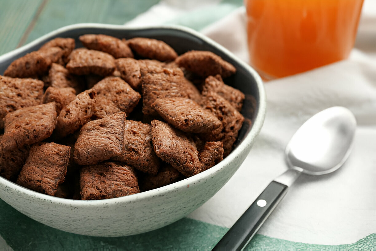 A bowl of chocolate-flavored cereal pieces with a silver spoon nearby and a glass of orange juice in the background.