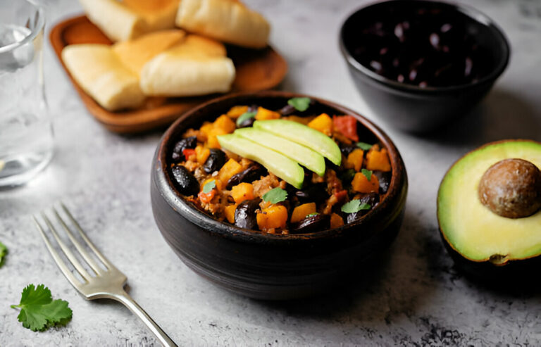 A bowl of colorful bean stew topped with apple slices, surrounded by bread rolls, avocado, and black beans.