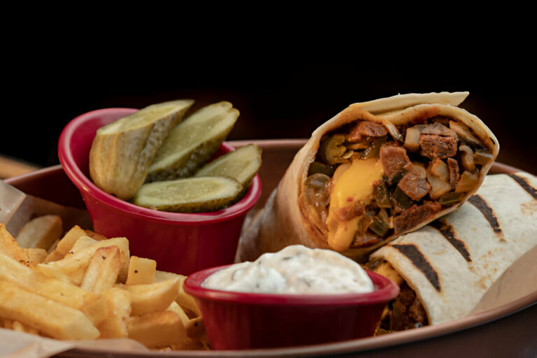 A delicious burrito with cheese, beef, and peppers, served with fries, pickles, and a creamy dip on a rustic plate.